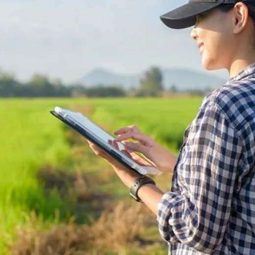 A woman wearing a baseball cap and flannel shirt stands in a field working on a tablet